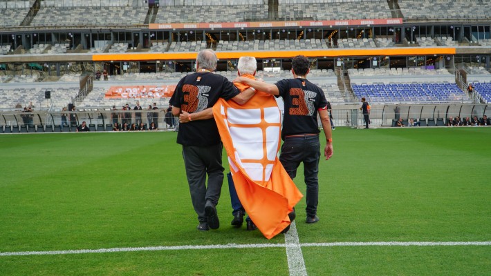 Três homens abraçados no gramado do estádio, com bandeira da Atex e camisetas com o número 35